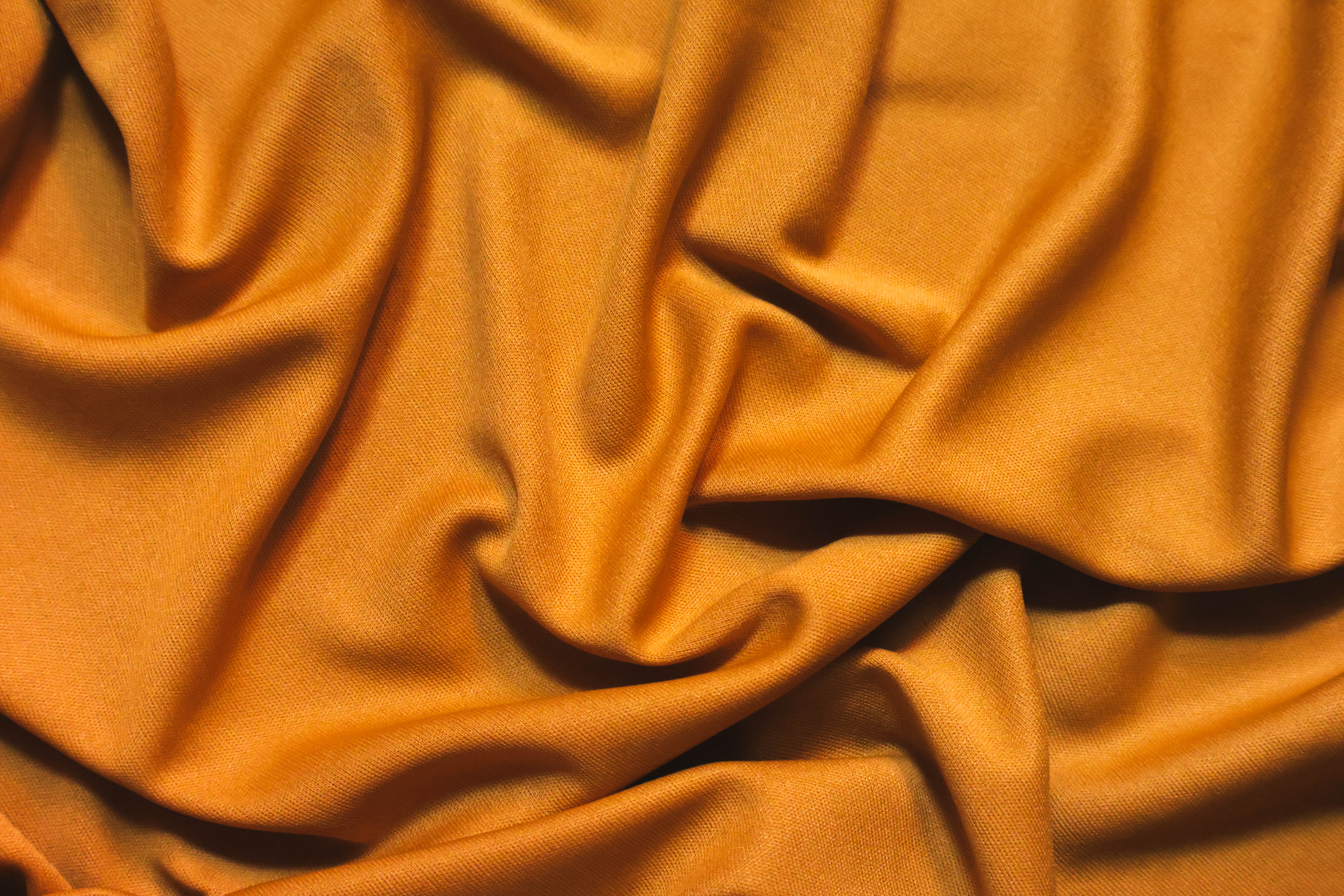 Fotografia. Close em tecido laranja com textura acetinada. O tecido está amassado, formando curvas e sombras suaves.