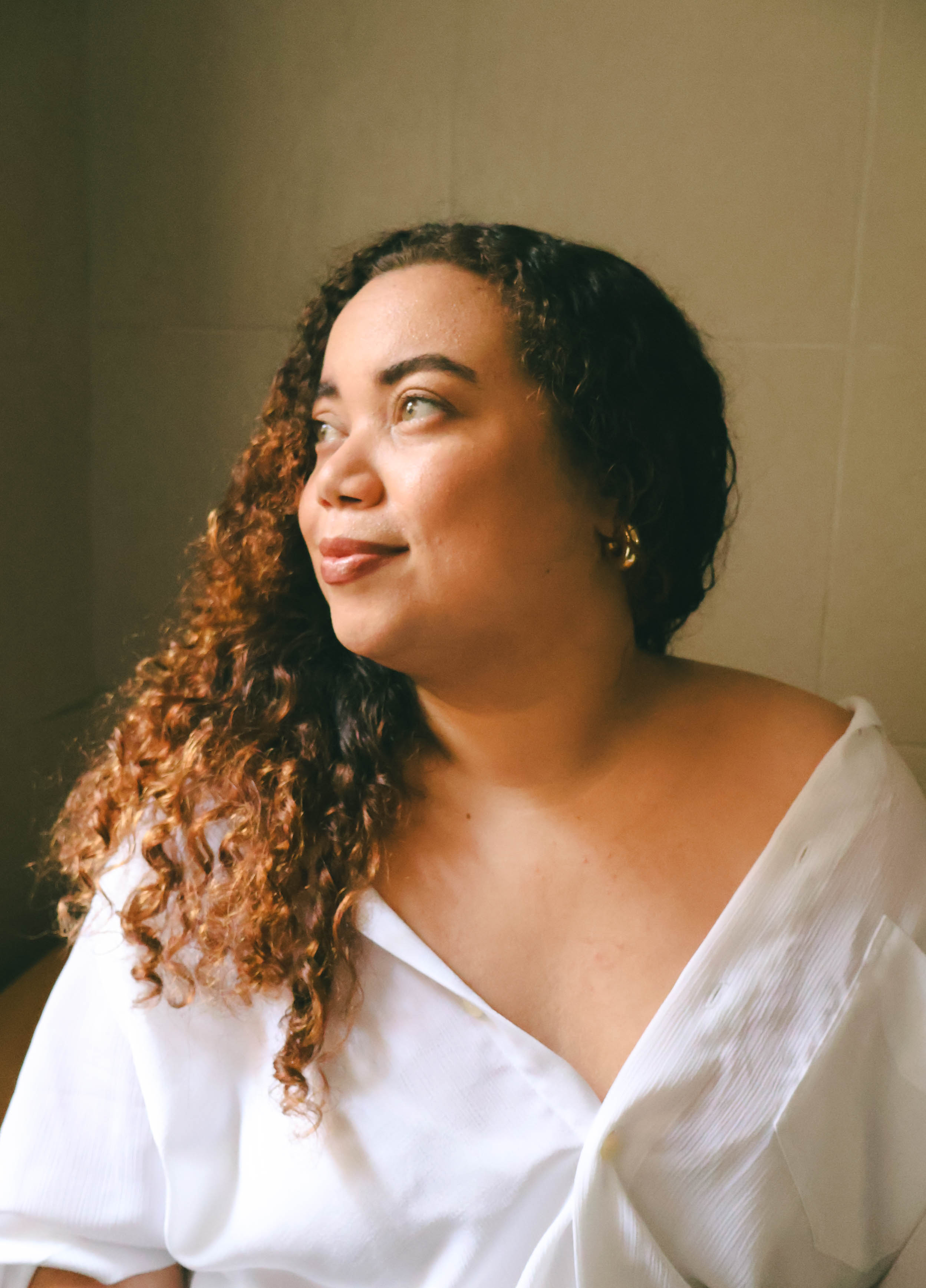Fotografia da artista. Mulher de pele clara, olhos verdes, rosto redondo, cabelos cacheados loiros-acizentados, usando uma camisa branca com um dos ombros à mostra. Está sentada, olhando levemente para o lado esquerdo da imagem com expressão serena. A luz natural entra pela lateral, iluminando suavemente seu rosto. O fundo é neutro, em tom bege claro.