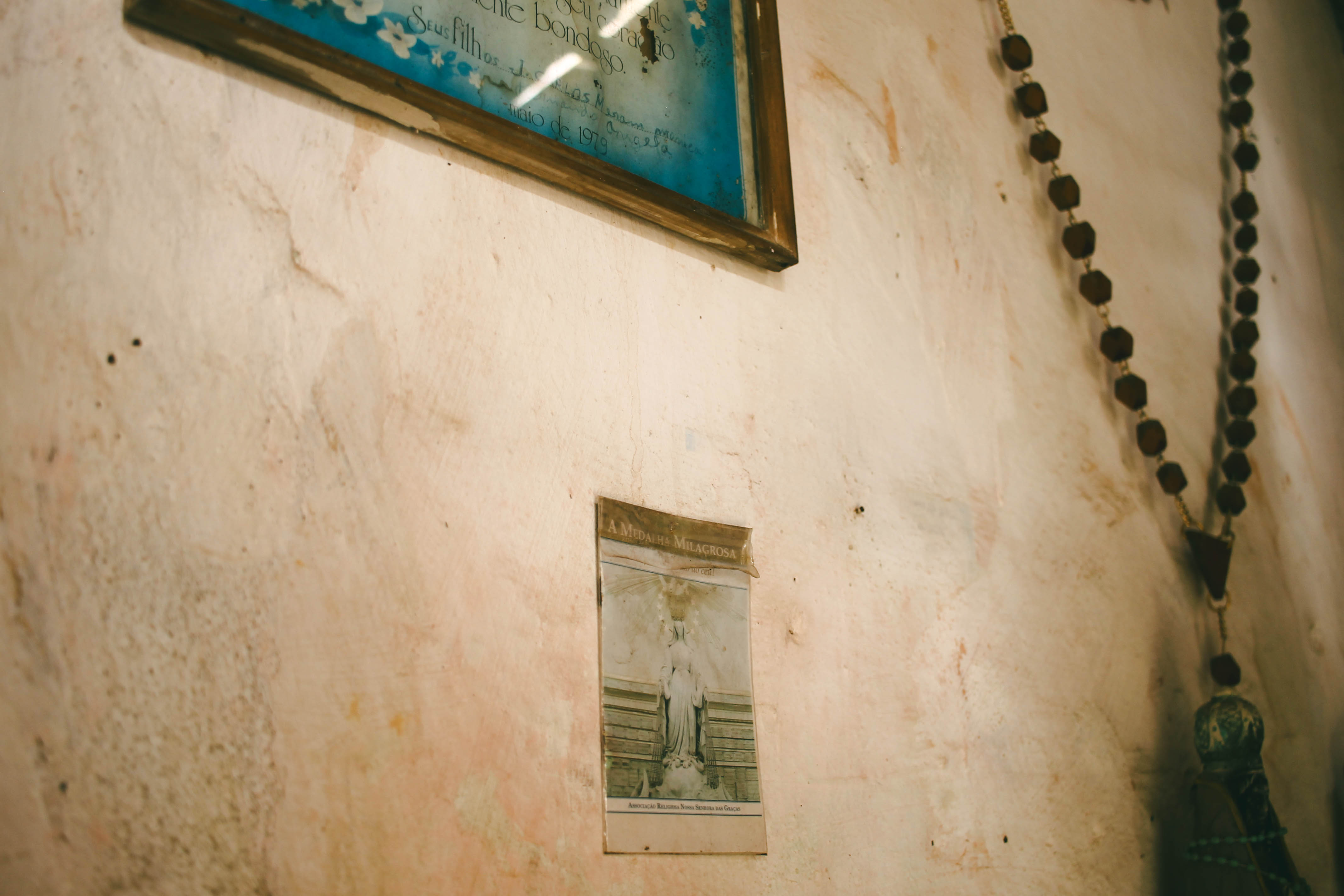 Fotografia. Parede de reboco com marcas de tempo. Há um terço pendurado, um quadro antigo com uma impressão em papel azul e um pedaço de papel colado ao lado.